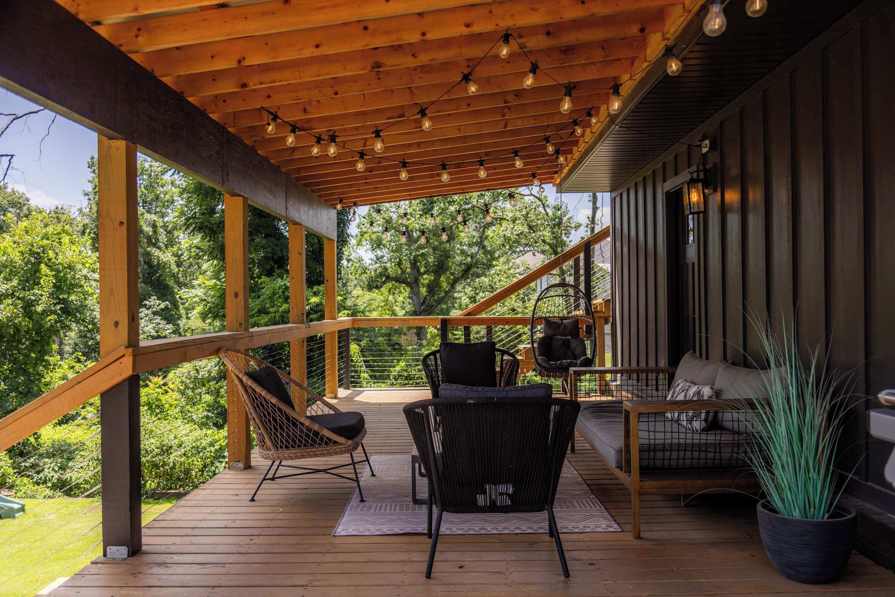 A wooden deck with outdoor seating, including chairs and a sofa, under a wooden pergola with string lights.
