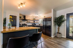 Une cuisine moderne avec un îlot central en bois, des chaises noires, un réfrigérateur en acier inoxydable et une plante en pot.