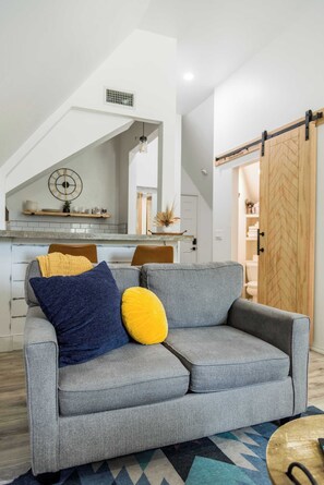 A modern living room with a grey sofa, a wooden door, and a kitchen area in the background. - The Loft | Modern Studio 5 Min to Downtown (Bentonville)