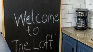 Quadro-negro com a inscrição 'Welcome to The Loft' escrita em giz branco, uma cafeteira sobre a bancada e um revestimento de azulejos na parede.