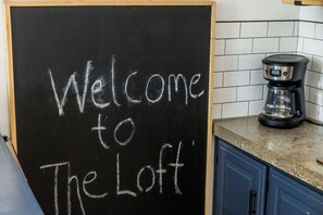Eine Tafel mit der Aufschrift 'Welcome to The Loft' in weißer Kreide, eine Kaffeemaschine auf der Arbeitsplatte und eine geflieste Spritzschutzleiste.
