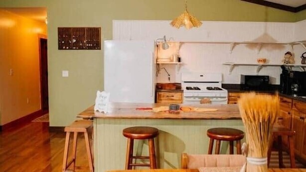 A dining area with a wooden table and chairs, a kitchen in the background, and a chandelier.