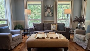 A living room with a brown leather sofa, two armchairs, a coffee table, and a fireplace. There are two lamps on side tables, a framed picture on the wall, and a window with curtains. - 5BR near Park & Downtown Bar Harbor! [North Lodge] (Bar Harbor)