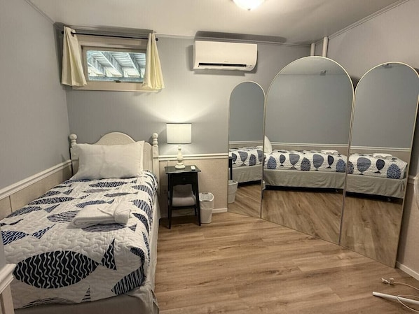 A bedroom with a bed, bedside table, lamp, air conditioning unit, window, and a mirror.