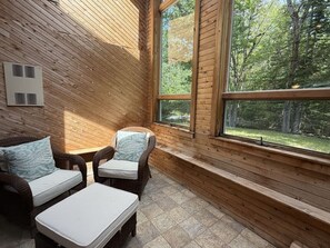 Ein Zimmer mit Holzwandverkleidung, zwei Korbstühlen und einem kleinen Tisch, ein Fenster mit Blick auf Bäume und einer Wandheizung.
