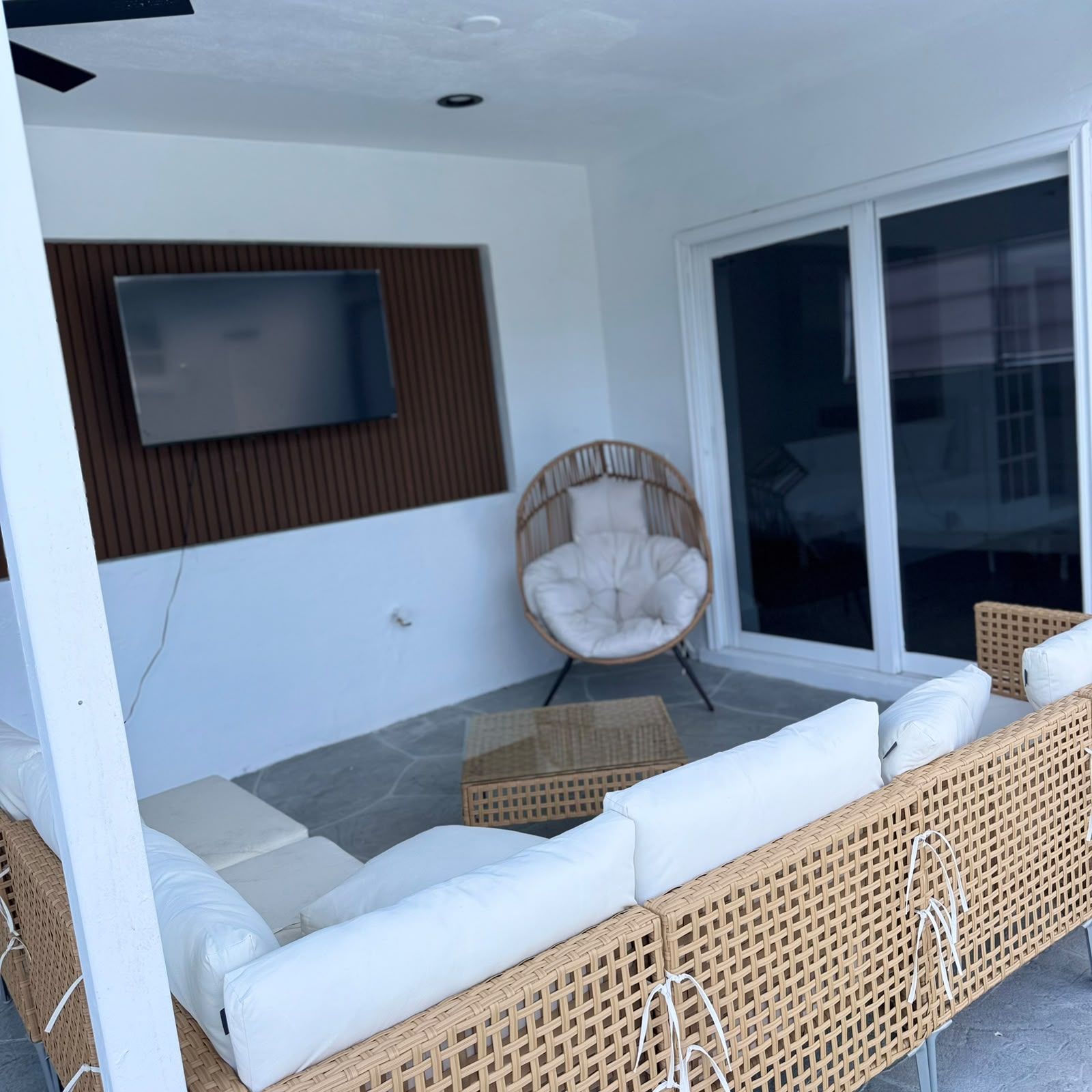Un espace de détente extérieur moderne, comprenant un canapé, un fauteuil en osier et un téléviseur à écran plat fixé au mur.