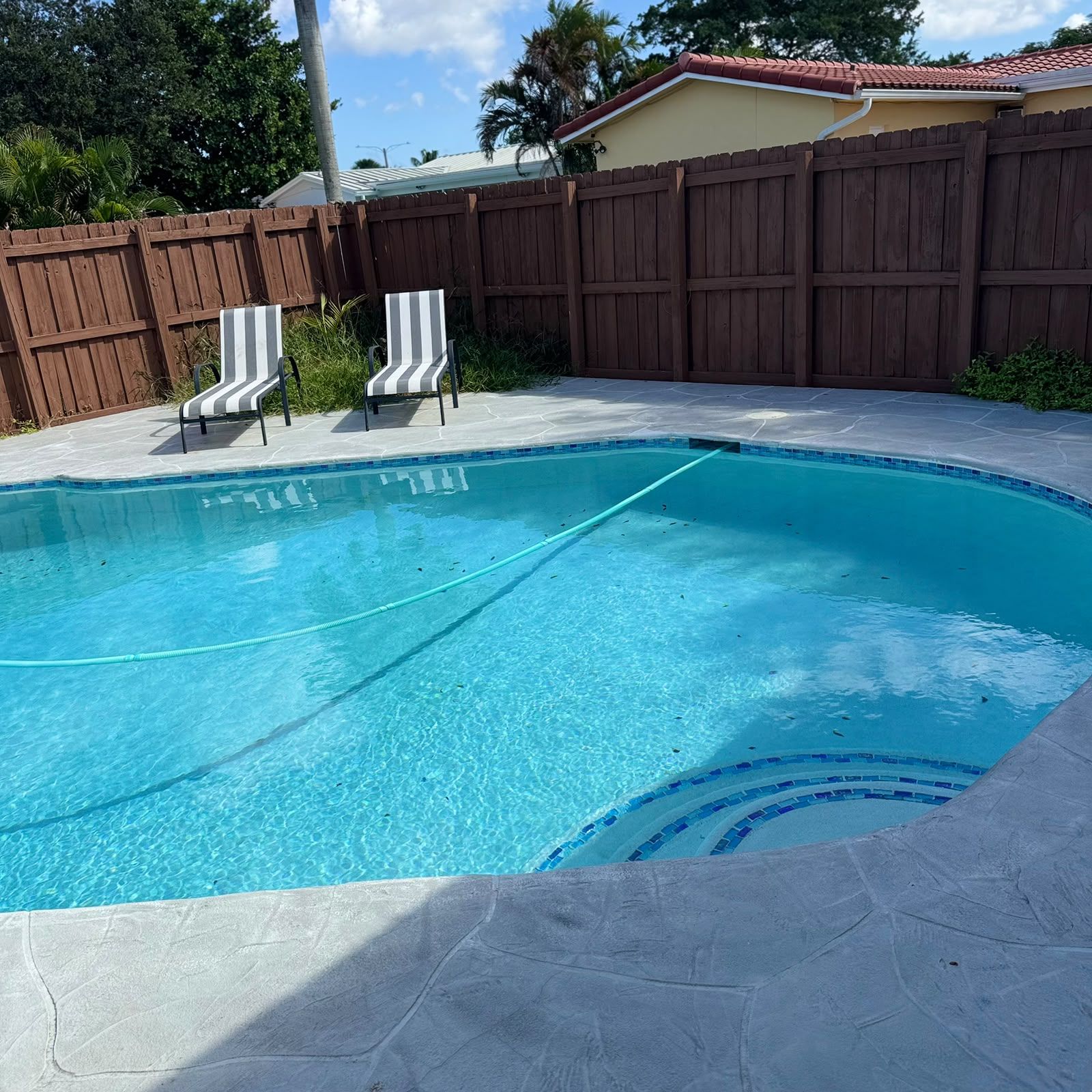 Une piscine entourée d’une clôture en bois, deux chaises longues à rayures et une terrasse carrelée.
