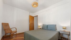 Una habitación con cama, una silla, una mesita de noche con lámpara y una puerta.