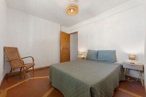 Una habitación con cama, una silla, una mesita de noche con lámpara y una puerta.
