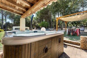 An outdoor hot tub under a wooden pergola with a pool and lounge chairs in the background.