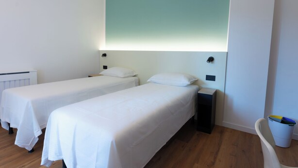 Una camera d'albergo con due letti singoli, un comodino con una lampada, una luce a parete e un termosifone.