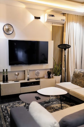 A modern living room with a flat-screen TV, a sofa, a coffee table, and a decorative plant. - Leemas 2 Bedroom Duplex  (Jos)