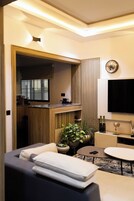 Uma sala de estar moderna com sofá, mesa de centro, TV e área de cozinha visível através de uma porta aberta.