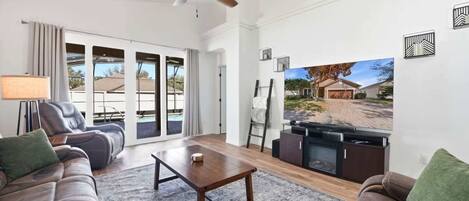 Un salón amplio con ventilador de techo, un televisor de pantalla plana grande y vistas a una zona residencial.