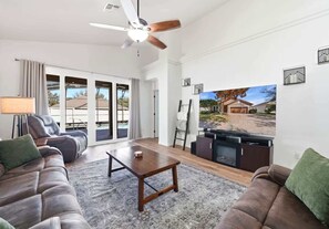 A spacious living room with a ceiling fan, a large flat-screen TV, and a view of a residential area. - Bridgeford Splash Retreat (Davenport)