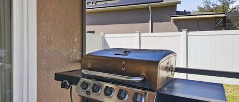 A stainless steel barbecue grill with four burners, positioned on a balcony with a view of a residential area and a white fence.