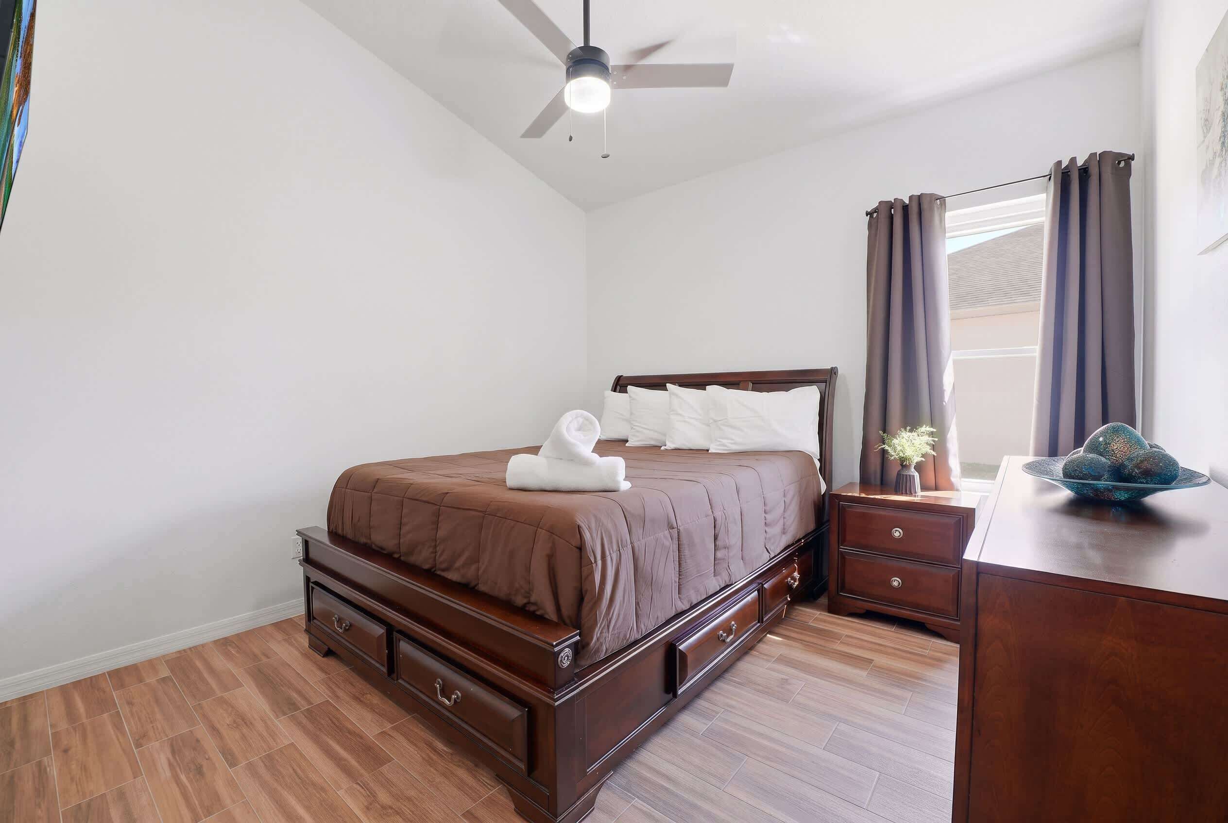 Un dormitorio con cama, cómoda, mesita de noche, ventilador de techo y ventana con cortinas.