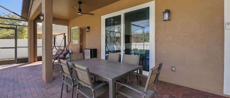 Un patio al aire libre con una mesa y sillas, un ventilador de techo y un ventanal de vidrio que da a una zona cubierta.
