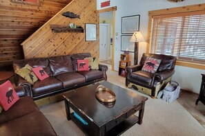Ein gemütliches Wohnzimmer mit einem braunen Ledersofa, einem passenden Sessel und einem Couchtisch. Im Hintergrund ist eine Holztreppe mit Fischdekor zu sehen.