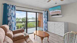 A living room with a sofa, a wooden coffee table, a TV mounted on the wall, and a balcony with a view of greenery. - PCB Beach Home With Gulf Access, Pool, and More (Panama City Beach)