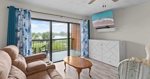 Ein Wohnzimmer mit einer Couch, einem Holztisch, einem an der Wand befestigten Fernseher und einem Balkon mit Blick auf Grünflächen.