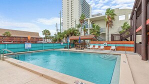 A swimming pool with lounge chairs and a covered seating area, surrounded by a fence and buildings. - PCB Beach Home With Gulf Access, Pool, and More (Panama City Beach)