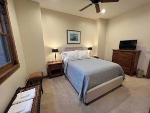 Quarto com cama, mesas de cabeceira, cômoda, ventilador de teto e janela.