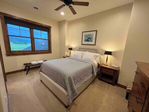 Une chambre à coucher comprenant un lit, des tables de chevet, un banc, une commode, un ventilateur de plafond et une fenêtre donnant sur un beau paysage.