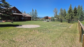 A grassy backyard with a wooden fence, a gravel area, and two buildings with red roofs. - Grizzly Home on Ski Hill Road, Targhee shuttle (Driggs)