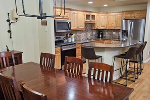 A modern kitchen with wooden cabinets, a stainless steel refrigerator, a microwave, and a central island with bar stools. - Mountain Town Getaway on Teton Creek (Driggs)