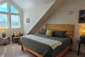 Ein Schlafzimmer mit schrägverdeckter Decke, einem Bett mit Holz Kopfteil, zwei Nachttischlampen, einem Sessel und einem Fenster mit Jalousien.