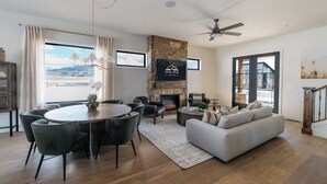 Een ruime woonkamer met een open haard, een eetgedeelte met een ronde tafel, en een groot raam met uitzicht op het omliggende landschap.