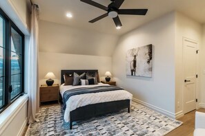 A bedroom with a bed, two bedside tables, a ceiling fan, a window with curtains, a painting on the wall, and a door. - White Pine Lodge in Teton Valley (Driggs)