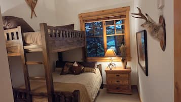 A cozy bedroom with a bunk bed, a bedside table with a lamp, a window with a view of trees, and a mounted deer head on the wall.