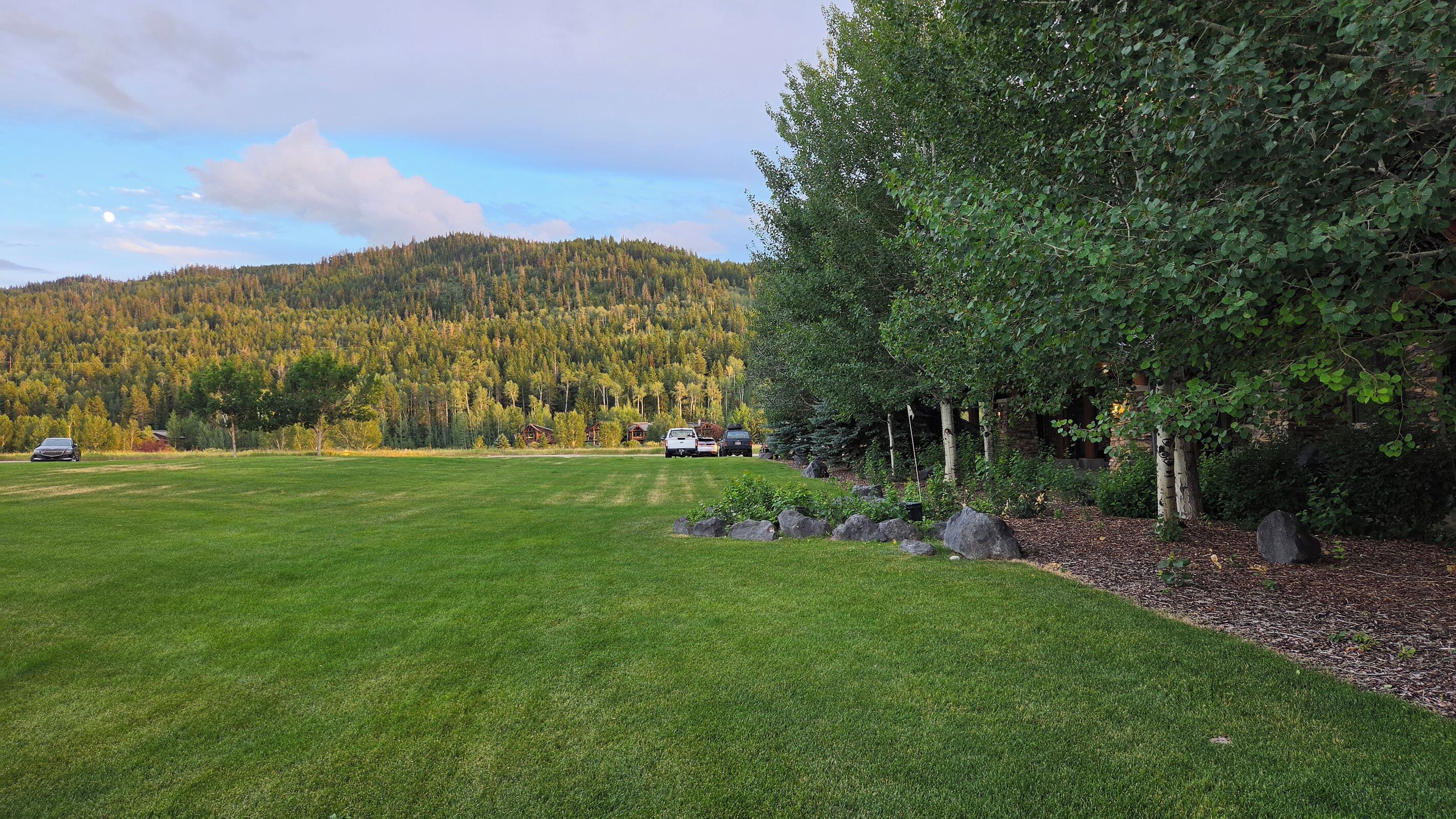A grassy field with a forested hill in the background and a building with a stone facade.