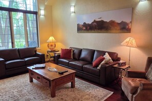 A living room with a brown leather sofa, a wooden coffee table, and a rug. There is a painting on the wall and two lamps on the side tables. - Teton Creek Getaway with Hot Tub (Driggs)