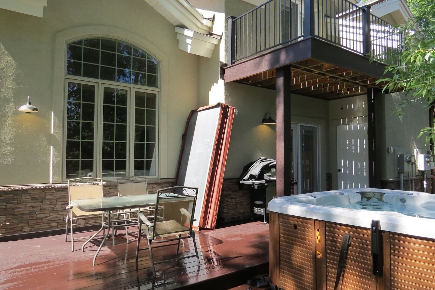 Un bain à remous sur une terrasse en bois, avec une table et des chaises en verre, une grande fenêtre et une porte dotée d’un portail en métal.