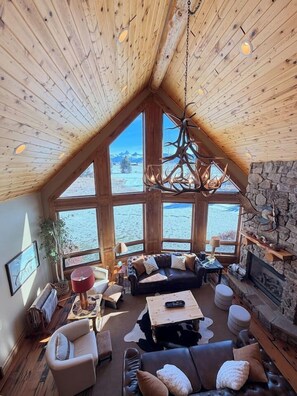 A spacious living area with a high ceiling, large windows, a stone fireplace, and a wooden ceiling. - It's Always "Peak" Season in the Tetons (Tetonia)