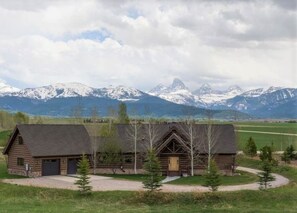 A rustic log cabin with a garage, surrounded by a grassy area and trees, with mountains in the background. - It's Always "Peak" Season in the Tetons (Tetonia)