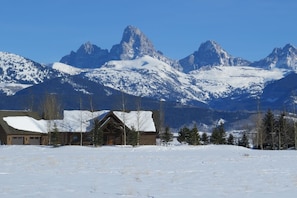 A cabin with a snow-covered roof, surrounded by snow-covered ground and trees, with mountains in the background. - It's Always "Peak" Season in the Tetons (Tetonia)