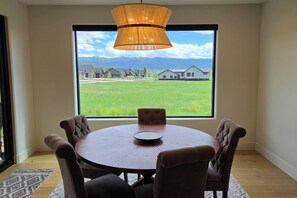 A dining area with a round table and six chairs, a large window with a view of a green field and houses, and a hanging light fixture. - Teton Reserve Royal Elk Lodge (Victor)