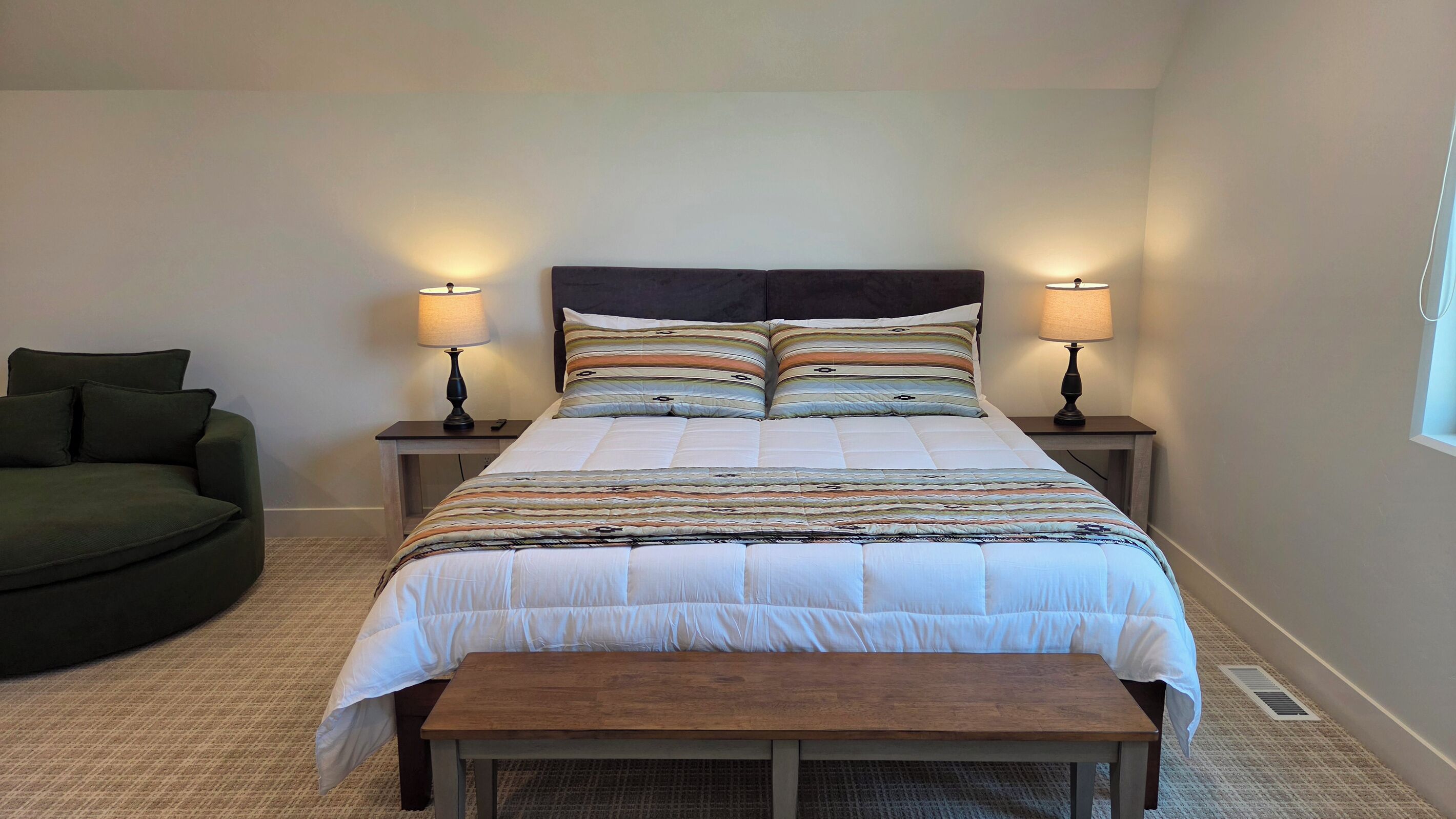 Een slaapkamer met een bed, twee nachtkastjes met lampen, een bankje en een groene fauteuil.