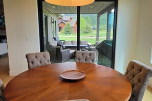 Ein runder Holztisch mit vier gepolsterten Stühlen, eine Glastrennwand und Blick auf eine Außenpatio mit Sitzgelegenheiten und Grillplatz.