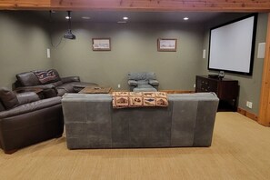 A living room with a grey sectional sofa, a brown leather couch, and a grey recliner. There is a projector mounted on the ceiling, a flat-screen TV on the wall, and two framed pictures on the wall.