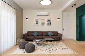 A modern living room with a brown sofa, two round poufs, a coffee table, and a patterned rug. - Modern 2BR with Sunny Balcony by HolyGuest (Tel Aviv-Yafo)