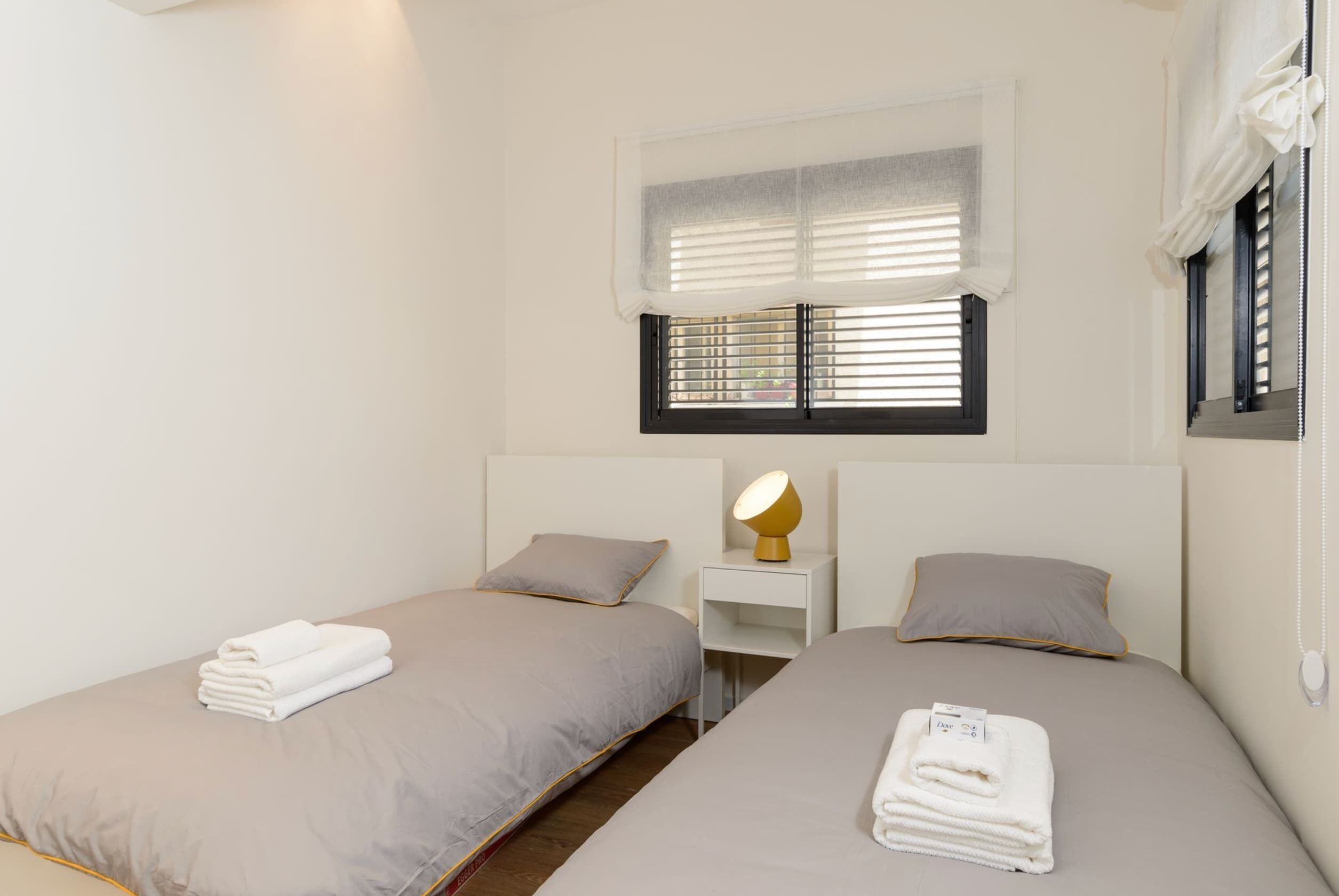 A hotel room with two beds, a nightstand with a lamp, a window with blinds, and a curtain.