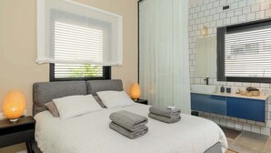 Une chambre moderne avec un grand lit, des lampes de chevet, une salle de bain avec un miroir et une vue sur la verdure à l’extérieur.