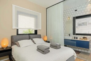 A modern bedroom with a large bed, bedside lamps, a bathroom with a mirror, and a view of greenery outside. - Modern 2BR with Sunny Balcony by HolyGuest (Tel Aviv-Yafo)