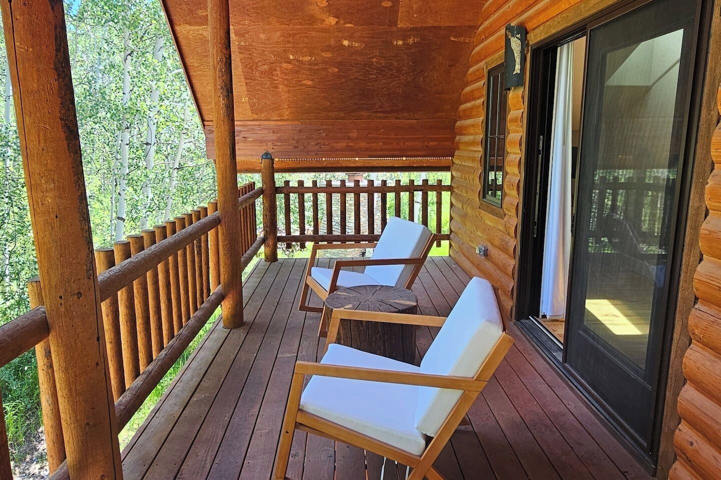 A wooden cabin with a covered porch, two white cushioned chairs, and a glass door leading to the interior.