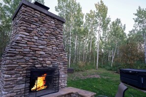 A stone fireplace with a fire burning inside, surrounded by a grassy area and trees.
